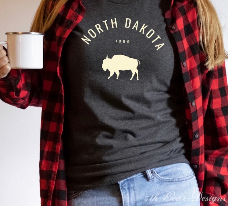 Puede incluir: Una camiseta gris oscura con un gr&aacute;fico blanco de un bisonte y el texto "NORTH DAKOTA 1889" en un c&iacute;rculo blanco.