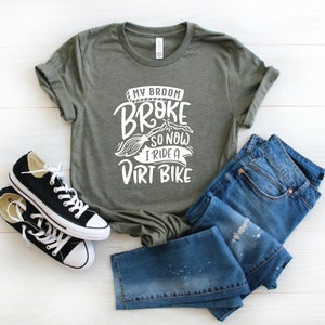 May include: A green t-shirt with white text that reads "My broom broke so now I ride a dirt bike". The shirt is paired with blue jeans and black sneakers.