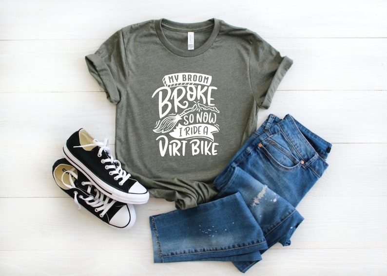 May include: A green t-shirt with white text that reads "My broom broke so now I ride a dirt bike". The shirt is paired with blue jeans and black sneakers.