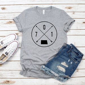 May include: Gray t-shirt with a black circle graphic featuring the numbers 071 and a black silhouette of the state of Kansas. The graphic also includes two arrows pointing in opposite directions.