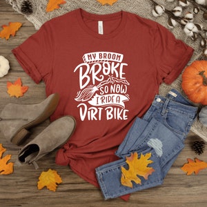 May include: A red t-shirt with white text that reads "My broom broke so now I ride a dirt bike". The shirt is laid out on a wooden surface with fall-themed decorations, including leaves, pumpkins, and a pair of brown ankle boots.