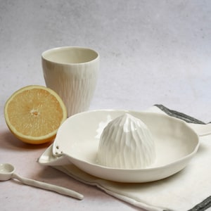 Op de afbeelding: Een witte keramische citruspers met een getextureerde ruimer, een bijpassende beker en een kleine lepel. Een halve grapefruit en een wit linnen servet met een grijze rand maken de scène compleet.