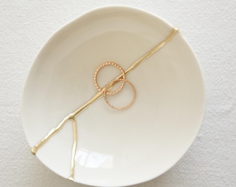 Puede incluir: Un plato de cerámica blanco con un diseño dorado en forma de grieta. Dos anillos dorados descansan sobre el plato. El plato tiene una forma orgánica y se encuentra sobre una superficie blanca texturizada. La estética general es minimalista y elegante.