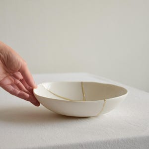 May include: A shallow, off-white ceramic bowl featuring a smooth, rounded silhouette. The exterior surface is accented with thin, gold-colored lines that appear to mend or highlight cracks across the curved body of the vessel.