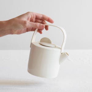 Puede incluir: Una tetera de cerámica blanca con un asa curva y un pequeño pico. La tetera tiene un cuerpo redondeado y una tapa con un asa pequeña y plana. La tetera está sostenida por una mano sobre un fondo blanco.
