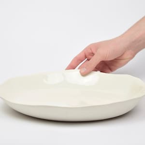 Op de afbeelding: Een witte keramische serveerschaal met een organische, golvende rand. De schaal is leeg en lijkt van een dik, duurzaam materiaal te zijn gemaakt.