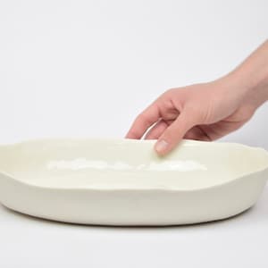 Könnte beinhalten: Eine weiße Keramik-Servierplatte mit leicht gewelltem Rand. Die Platte ist oval geformt und hat eine glatte, glänzende Oberfläche.