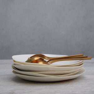 Pode incluir: Uma pilha de pratos brancos sujos, com formato único, com um conjunto de colheres douradas em cima. Os pratos têm uma forma suave e orgânica, e as colheres têm um design elegante e moderno. O fundo é cinza neutro.