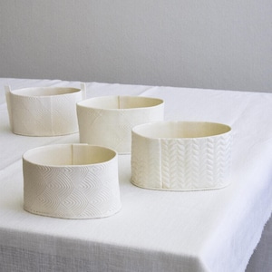 May include: Four off-white ceramic containers with textured patterns. The containers are cylindrical with varying heights and designs, including chevron and leaf-like patterns. They are arranged on a white surface.