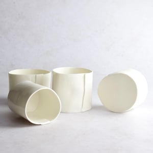 May include: Four white ceramic cups, three are upright and one is laying on its side. The cups have a slightly textured surface.