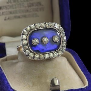 Op de afbeelding: Een vintage ring met een rechthoekige blauwe middensteen, geaccentueerd door drie kleinere diamanten. De ring is omgeven door een halo van kleine diamanten en gezet in een zilveren of witgouden zetting. Gepresenteerd in een blauwe fluwelen ringdoos.
