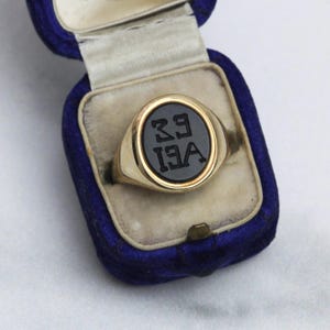May include: A gold signet ring with a black oval center featuring the letters "39" over "13A". The ring is displayed in a blue velvet ring box with a white interior. The ring has a polished, shiny finish.