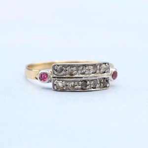 May include: A gold ring with a rectangular silver setting. The setting is adorned with two rows of small, round, clear stones. Small, round, red stones are set on each side of the rectangular setting. The ring has a delicate, vintage appearance.