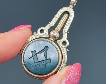 May include: A gold-coloured pendant with a dark green circular centre featuring a Masonic symbol. The pendant has an ornate design with a hinged top and a decorative bail. The symbol includes a square, compass, and ruler.