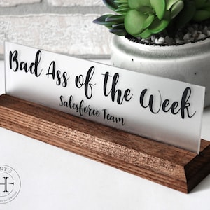 May include: A frosted acrylic desk sign with the text "Bad Ass of the Week Salesforce Team" on a dark brown wooden base.