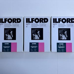 Peut inclure: Trois bo&icirc;tes de papier photographique Ilford sont pr&eacute;sent&eacute;es c&ocirc;te &agrave; c&ocirc;te. Chaque bo&icirc;te affiche le logo Ilford en noir, une photographie en noir et blanc et un accent rectangulaire rose. Les bo&icirc;tes sont &eacute;tiquet&eacute;es "Multigrade IV RC de Luxe".