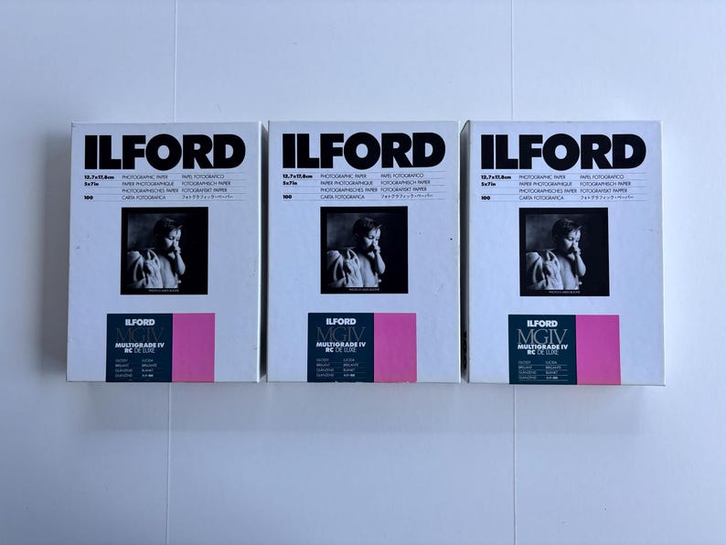 Peut inclure: Trois bo&icirc;tes de papier photographique Ilford sont pr&eacute;sent&eacute;es c&ocirc;te &agrave; c&ocirc;te. Chaque bo&icirc;te affiche le logo Ilford en noir, une photographie en noir et blanc et un accent rectangulaire rose. Les bo&icirc;tes sont &eacute;tiquet&eacute;es "Multigrade IV RC de Luxe".