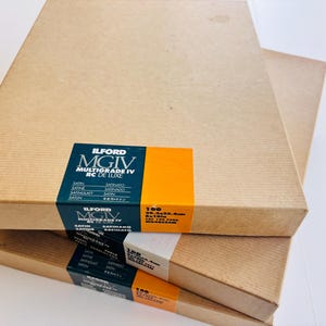 Peut inclure: Trois bo&icirc;tes rectangulaires empil&eacute;es de papier photo Ilford MGIV Multigrade IV RC de Luxe. Les bo&icirc;tes sont marron clair avec des &eacute;tiquettes noires et orange. Chaque bo&icirc;te contient 100 feuilles de papier 20,3 x 25,4 cm.