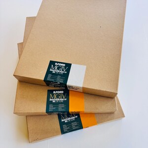 Peut inclure: Quatre bo&icirc;tes rectangulaires marron clair empil&eacute;es. Chaque bo&icirc;te porte une &eacute;tiquette noire, blanche et orange avec l'inscription "ILFORD MGIV MULTIGRADE IV RC DE LUXE". Les bo&icirc;tes sont en carton.