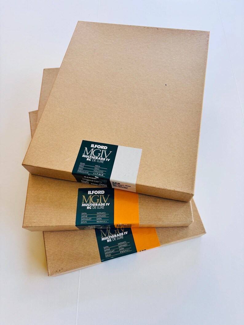 Peut inclure: Quatre bo&icirc;tes rectangulaires marron clair empil&eacute;es. Chaque bo&icirc;te porte une &eacute;tiquette noire, blanche et orange avec l'inscription "ILFORD MGIV MULTIGRADE IV RC DE LUXE". Les bo&icirc;tes sont en carton.