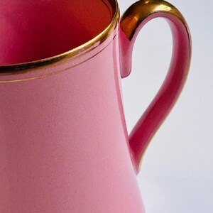 Pu&ograve; includere: Un'anfora in ceramica rosa con un bordo e un manico dorati. L'anfora ha una forma svasata e una finitura liscia e lucida.