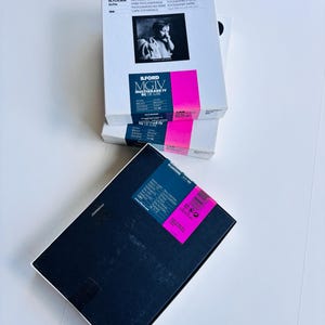 Peut inclure: Plusieurs bo&icirc;tes de papier photographique Ilford sont empil&eacute;es. Les bo&icirc;tes sont blanches avec du texte noir et une bande rose. La bo&icirc;te sup&eacute;rieure pr&eacute;sente une photo en noir et blanc. Le texte "ILFORD MGIV MULTIGRADE IV RC DE LUXE" est visible.