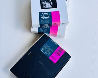 May include: A stack of Ilford photographic paper boxes. The boxes are white with black text and a pink stripe. The top box features a black and white photograph. The text "ILFORD MGIV MULTIGRADE IV RC DE LUXE" is visible on the boxes. The boxes are on a white surface.