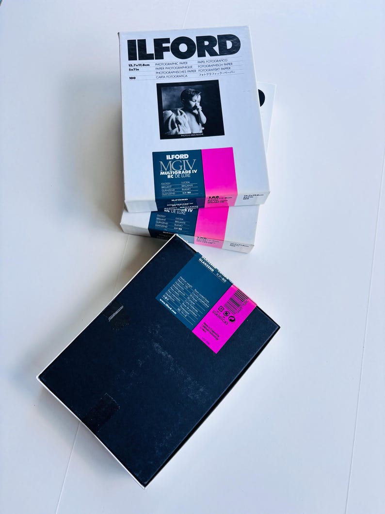 Peut inclure: Plusieurs bo&icirc;tes de papier photographique Ilford sont empil&eacute;es. Les bo&icirc;tes sont blanches avec du texte noir et une bande rose. La bo&icirc;te sup&eacute;rieure pr&eacute;sente une photo en noir et blanc. Le texte "ILFORD MGIV MULTIGRADE IV RC DE LUXE" est visible.