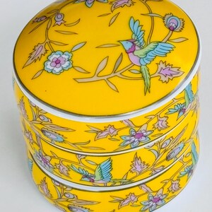 Peut inclure: Une boîte à bijoux en céramique jaune à trois niveaux avec un couvercle. La boîte est décorée d'un motif d'oiseaux et de fleurs colorées. L'oiseau a des plumes bleues, roses et vertes. Le motif floral comprend des fleurs roses et bleues.