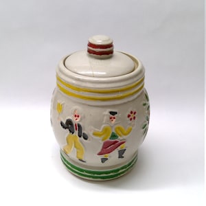 Op de afbeelding: Een witte keramische pot met een deksel, versierd met een volkskunstontwerp van twee dansende mensen. De pot heeft groene, gele en rode accenten.