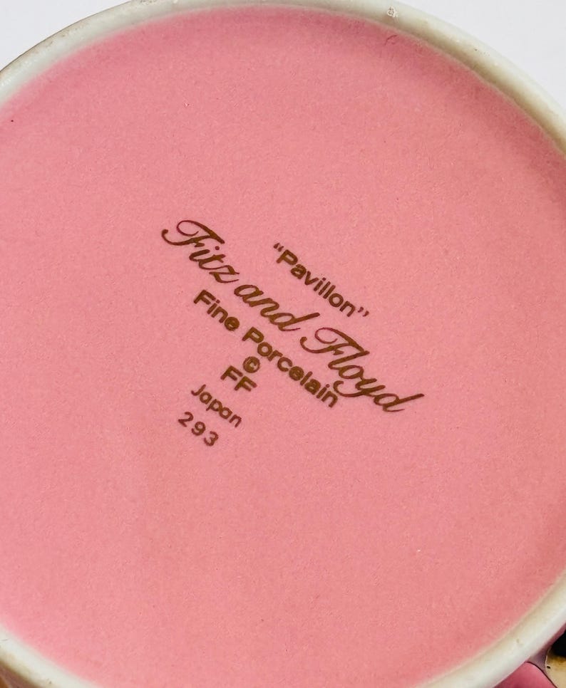 Pu&ograve; includere: Primo piano del fondo di una tazza in ceramica rosa con il testo "Fitz and Floyd" in lettere marroni. Il testo "Pavillon" &egrave; anche in lettere marroni. Il testo "Fine Porcelain" &egrave; in lettere marroni. Il testo "FF" &egrave; in lettere marroni. Il testo "Japan 293" &egrave; in lettere marroni.