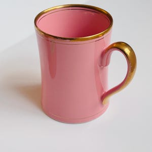 Pu&ograve; includere: Una tazza in ceramica rosa con un bordo e un manico dorati. La tazza &egrave; vuota e si trova su una superficie bianca.