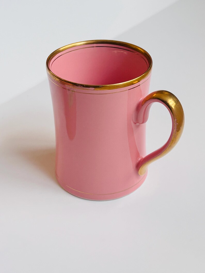 Pu&ograve; includere: Una tazza in ceramica rosa con un bordo e un manico dorati. La tazza &egrave; vuota e si trova su una superficie bianca.