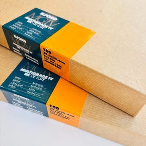 Peut inclure: Deux bo&icirc;tes rectangulaires de papier photo Ilford Multigrade IV RC de Luxe. Chaque bo&icirc;te a une &eacute;tiquette bleue et orange avec des informations sur le produit, dont la taille : 20,3 x 25,4 cm. Les bo&icirc;tes sont envelopp&eacute;es de papier brun.