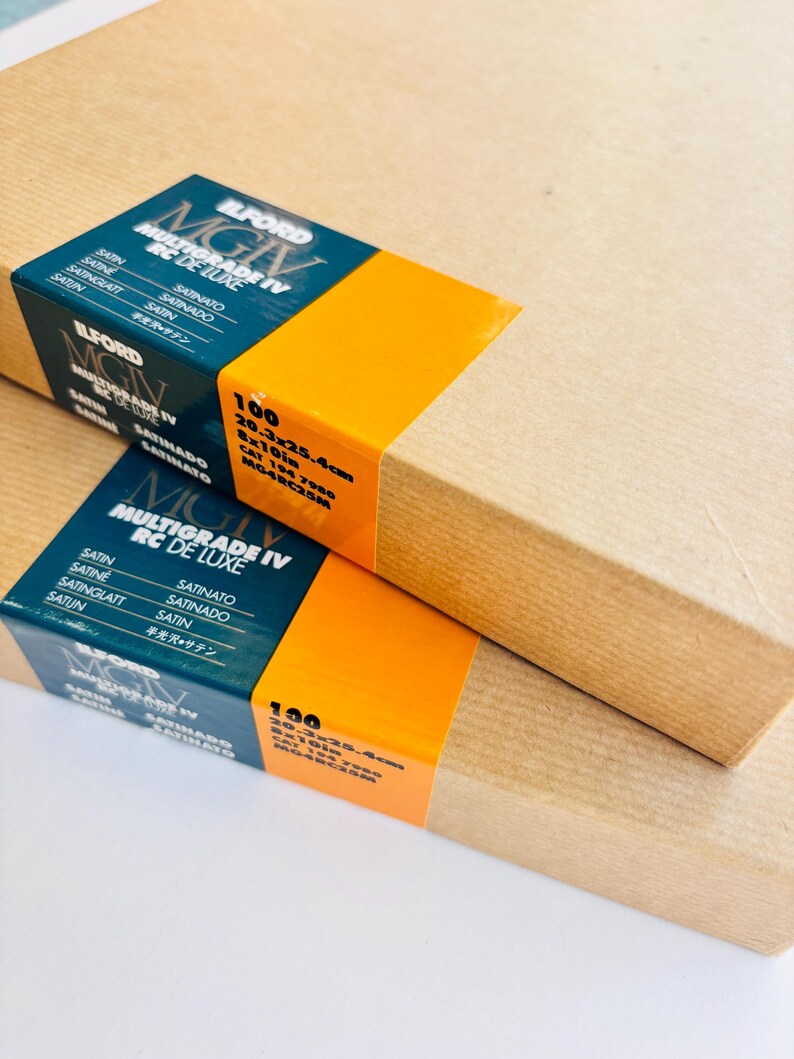 Peut inclure: Deux bo&icirc;tes rectangulaires de papier photo Ilford Multigrade IV RC de Luxe. Chaque bo&icirc;te a une &eacute;tiquette bleue et orange avec des informations sur le produit, dont la taille : 20,3 x 25,4 cm. Les bo&icirc;tes sont envelopp&eacute;es de papier brun.