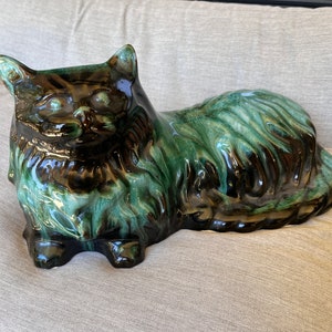May include: A ceramic cat figurine in a green and brown glaze. The cat is lying down with its head turned to the side. The glaze has a mottled effect, giving the cat a furry appearance.