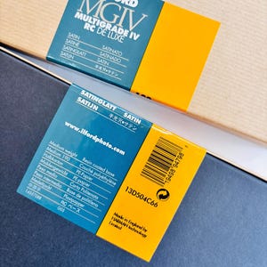 Peut inclure: Deux bo&icirc;tes de papier photographique Ilford MGIV Multigrade IV RC De Luxe. Les bo&icirc;tes sont bleu fonc&eacute; et jaune, avec du texte en plusieurs langues, dont "SATIN" et "SATINGLATT". Une bo&icirc;te porte un code-barres et le texte "Made in England by HARMAN technology Limited."