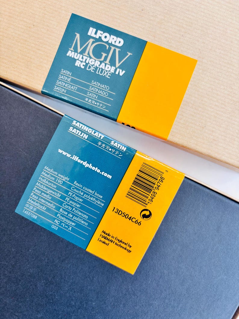 Peut inclure: Deux bo&icirc;tes de papier photographique Ilford MGIV Multigrade IV RC De Luxe. Les bo&icirc;tes sont bleu fonc&eacute; et jaune, avec du texte en plusieurs langues, dont "SATIN" et "SATINGLATT". Une bo&icirc;te porte un code-barres et le texte "Made in England by HARMAN technology Limited."