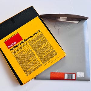 Peut inclure: Une bo&icirc;te Kodak jaune et noire avec le texte "feuilles adh&eacute;sives, type 2" et un rectangle rouge avec le logo Kodak. La bo&icirc;te est ouverte, r&eacute;v&eacute;lant une enveloppe grise.