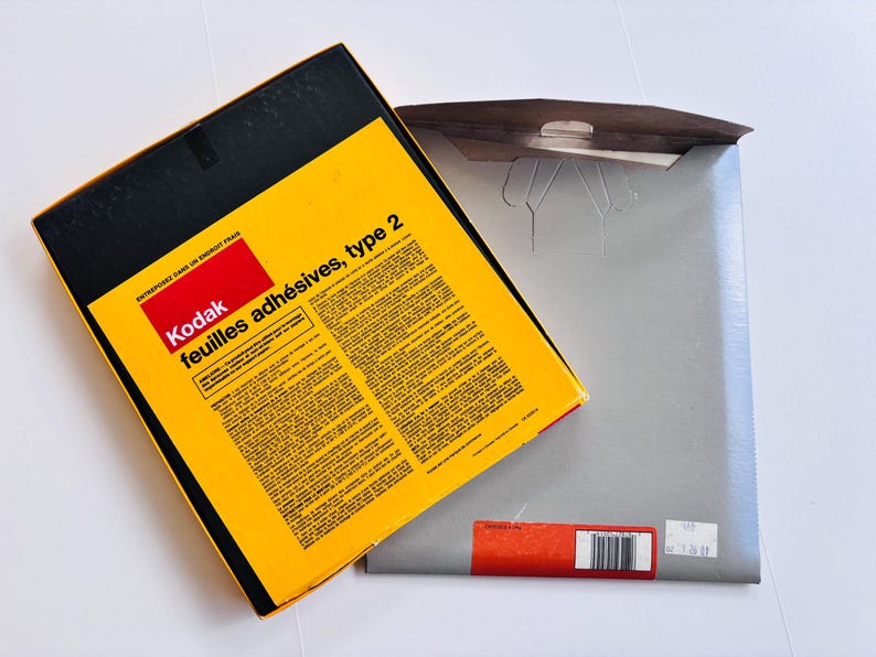 Peut inclure: Une bo&icirc;te Kodak jaune et noire avec le texte "feuilles adh&eacute;sives, type 2" et un rectangle rouge avec le logo Kodak. La bo&icirc;te est ouverte, r&eacute;v&eacute;lant une enveloppe grise.