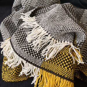 Peut inclure: Une couverture tissée avec des motifs géométriques noirs, blancs et jaunes. La couverture a des franges sur tous les côtés.