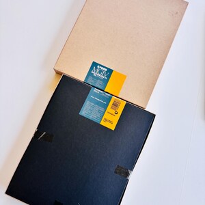Peut inclure: Deux bo&icirc;tes rectangulaires, une beige et une noire, sont empil&eacute;es sur une surface blanche. La bo&icirc;te noire porte une &eacute;tiquette bleue et jaune avec du texte. La bo&icirc;te beige est unie. Les deux bo&icirc;tes sont rectangulaires.