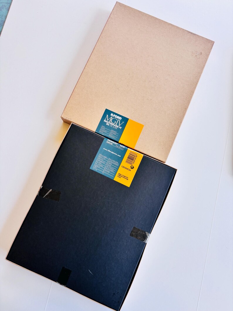 Peut inclure: Deux bo&icirc;tes rectangulaires, une beige et une noire, sont empil&eacute;es sur une surface blanche. La bo&icirc;te noire porte une &eacute;tiquette bleue et jaune avec du texte. La bo&icirc;te beige est unie. Les deux bo&icirc;tes sont rectangulaires.