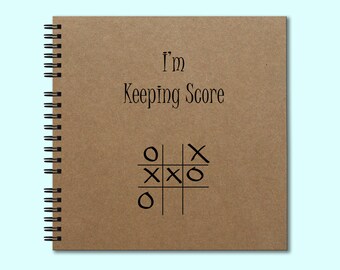 May include: Spiral-bound notebook with a kraft paper cover. The cover features the text "I'm Keeping Score" above a tic-tac-toe game in progress. The notebook has a black spiral binding.