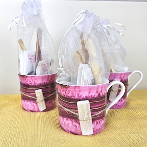 May include: Three pink mugs with white handles, each filled with a white organza bag containing wooden stirrers and small packets of hot chocolate mix. The mugs are decorated with gold and pink stripes and have a white crystal on the front.
