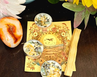 May include: Three small tealight candles with dried herbs and crystals inside, a stick of incense, and a carnelian crystal on a wooden surface with a vintage tarot card featuring a sun and birds.