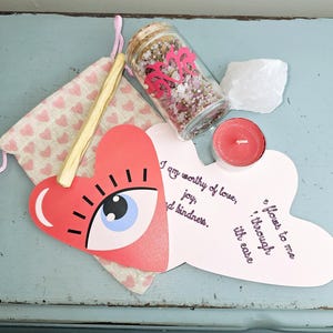 May include: A pink and white heart-shaped card with the text "I am worthy of love, joy, and kindness. Flowers to me through ease." A small glass jar filled with pink and white crystals, a pink candle, and a wooden stick are also in the image.