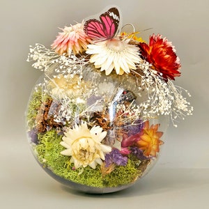 May include: A glass orb filled with dried flowers and greenery, including white, pink, red, and purple flowers, and green moss. A pink butterfly is perched on top of the arrangement.