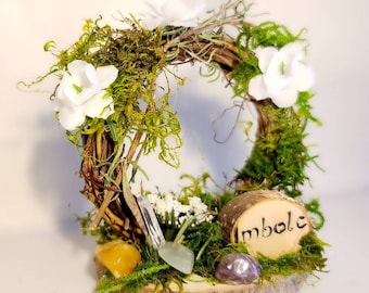 Puede incluir: Una pequeña corona de madera decorada con musgo, flores blancas y cristales. La corona está sentada sobre una rodaja de madera con la palabra "Imbolc" tallada en ella.