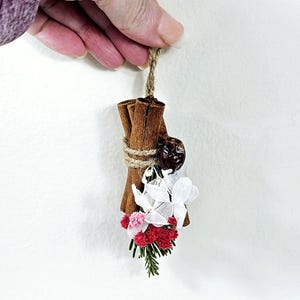 Puede incluir: Un manojo decorativo de palitos de canela atados con hilo, adornado con flores secas blancas, rosas y rojas, y una baya oscura. Cuelga de una cuerda de fibra natural, creando un objeto decorativo r&uacute;stico.